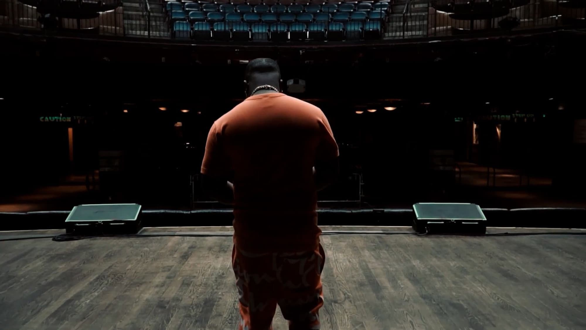 A man in an orange tracksuit stands alone on an empty theatre stage, facing the seats.